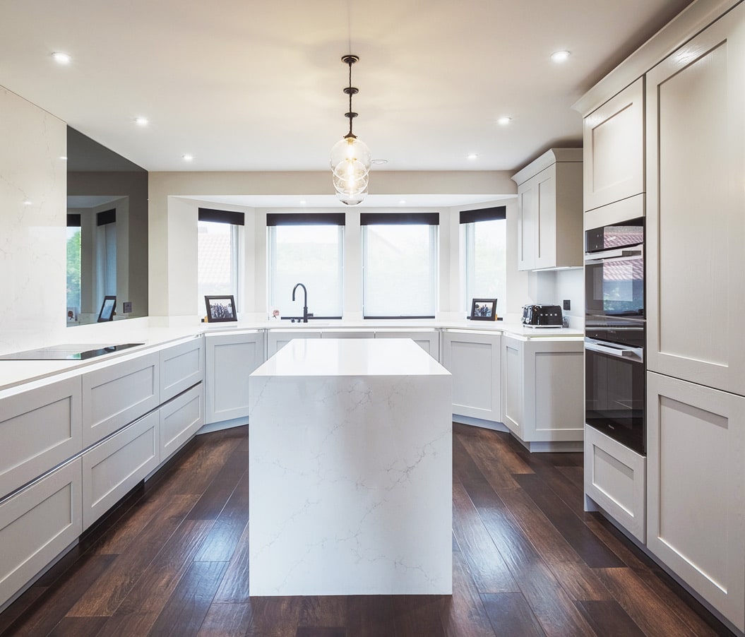 Modern white kitchen with island and dark wood floor, under bright ceiling lights.