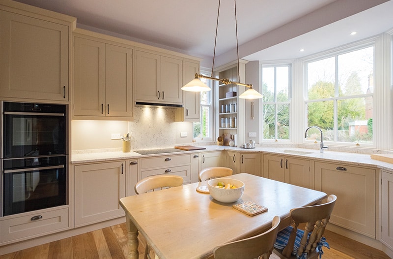 Bright kitchen with cream cabinets, a wooden table, and large windows letting in natural light.