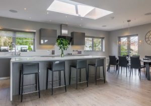 Modern kitchen with large island, black bar stools, dining area, and bright natural light from skylights.