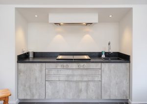 Modern kitchenette with grey cabinets, black countertop, and built-in stove, next to large windows.
