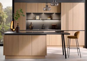 Modern kitchen with light wood cabinets, black countertop, and a single barstool by large windows.