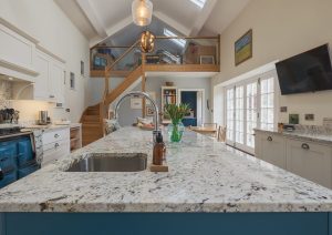 Modern kitchen with a marble island, sink, and blue accents, featuring natural light and wooden staircase.