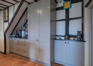 Gray kitchen cabinets and shelves beneath a staircase in a rustic-style room with tiled flooring.