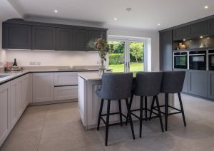 Modern kitchen with grey cabinets, island seating, built-in ovens, and large window showing greenery outside.