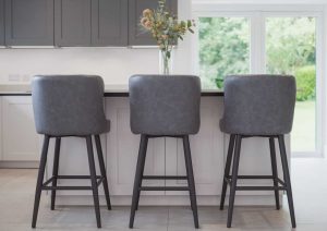 Three gray barstools at a kitchen island with a vase of flowers, near large windows.