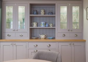 Light grey kitchen cabinet with glass doors, open shelves, and decorative mugs and teapots displayed.
