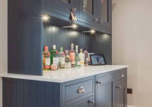 Home bar with bottles and glasses on a white countertop beneath blue cabinets with built-in lighting.
