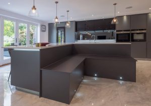 Modern kitchen with dark cabinets, built-in appliances, and bench seating with large windows in the background.